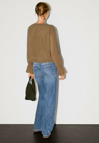 Braune semitransparente Bluse mit Rüschensäumen, kombiniert mit hellblauen weit geschnittenen Jeans. Das Model hält eine schwarze Handtasche.