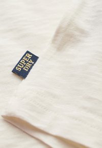 Hvid bomulds T-shirt med et struktureret stof. Har et marineblåt mærke med teksten "SUPER DRY" i guld, syet i kanten.