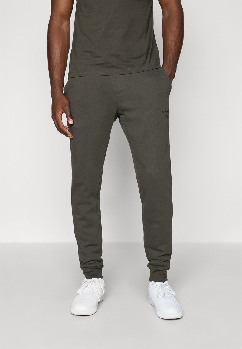Dark green cotton jogger pants with a slim fit, side pockets, and elastic cuffs. Pairing with white sneakers. Subtle logo detail on leg.