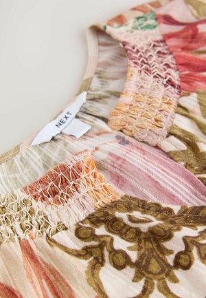 Prim-plan al unui material cu modele florale multicolore și detalii elastice smocate lângă o etichetă de marcă „NEXT” pe un fundal bej.