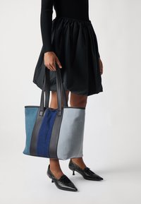 Sac fourre-tout en denim avec des accents en cuir noir. Présente des panneaux bleus, gris et marine. Deux anses noires. Tissu texturé.