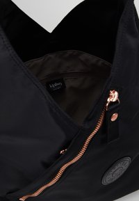 Zwarte nylon tote bag met een ritssluiting, rosegouden hardware en een rond logo-patch. Het interieur is voorzien van een zachte, donkere stoffen voering.