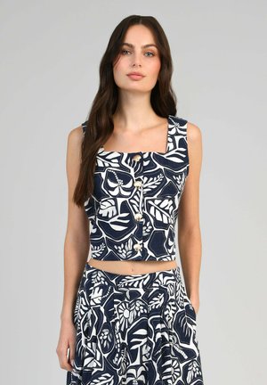 Femme portant un crop top sans manches à motif floral bleu marine et blanc avec des boutons et une jupe taille haute assortie, debout devant un fond gris uni.