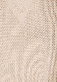 Sweter dzianinowy w jasnym beżowym kolorze z dekoltem w kształcie litery V. Tkanina ma teksturowany, luźny splot na całej powierzchni.