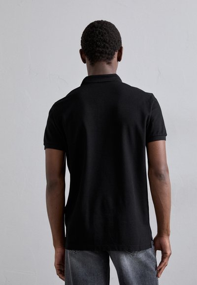 Schwarzes Polo-Shirt aus weichem Stoff, mit kurzen Ärmeln und klassischem Kragen, von hinten betrachtet. Getragen mit grauen Jeans.
