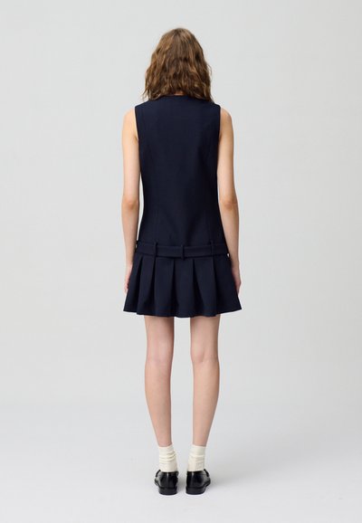 Robe sans manches bleu marine avec une jupe plissée, un design structuré et des liens au dos. Portée avec des chaussures noires et des chaussettes blanches.