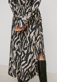 Schwarz-beiges gemustertes Kleid mit langen Ärmeln und einem seitlichen Schlitz, aus glattem Stoff. Die Hände sind auf der Taille positioniert.