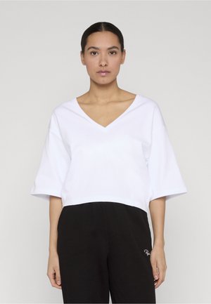 Kobieta o ciemnych włosach związanych z tyłu, ubrana w luźną białą bluzkę z dekoltem w kształcie litery V i szerokimi rękawami oraz czarne, teksturowane spodnie, stoi na tle jednolitego tła.