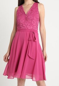 Rosa klänning med spetsliv och V-ringning, med en figursydd midja och flödande kjol. Inkluderar en knuten sashes detalj vid midjan.