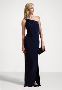 Marinblå one-shoulder klänning i mjukt tyg, med en sidofyra och samlad detalj vid axeln; i kombination med en svart clutch och klacksandaler.