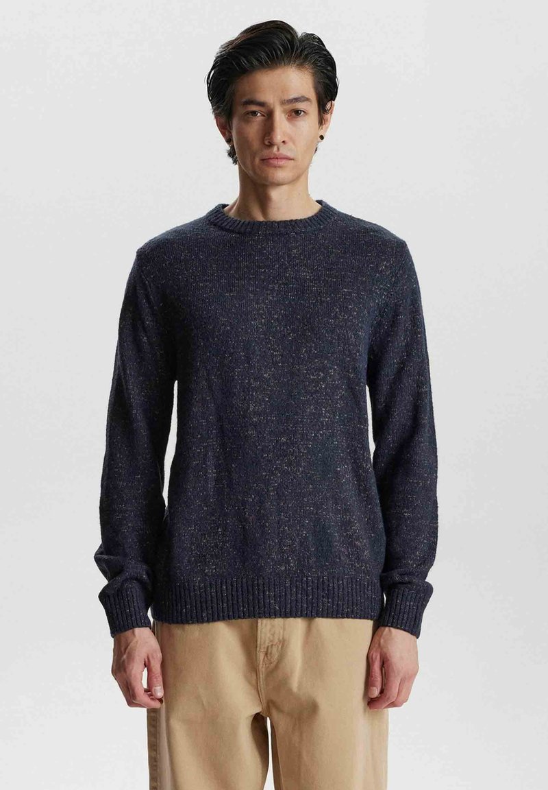 Granatowy sweter dziany z okrągłym dekoltem, teksturowanym materiałem i ściągaczami. Noszony z beżowymi spodniami, prezentując luźny krój i swobodny styl.