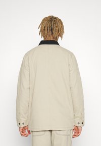 Beige Jacke mit schwarzem Kragen, locker geschnitten, mit geknöpften Ärmeln und sichtbaren Nähten. Der Rücken ist schlicht und glatt.