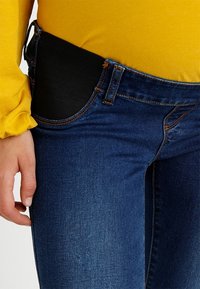 Tunna blå denimjeans med en svart elastisk panel vid midjan. Ovanpå är en långärmad gul skjorta.