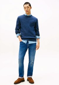 Marineblauer gesprenkelter Pullover über einem hellblauen Hemd, kombiniert mit ausgewaschenen blauen Jeans und braunen Wildlederschuhen. Das Model steht mit den Händen in den Taschen.