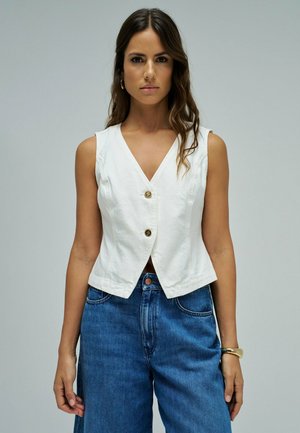 Femme aux longs cheveux ondulés portant un gilet blanc boutonné sans manches et un jean bleu à jambes larges, debout devant un fond gris uni.