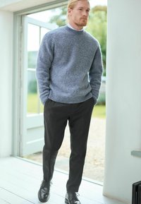 Pull en laine texturé gris avec un col rond, associé à un pantalon noir bien taillé et des chaussures de ville noires, se tenant près d'une fenêtre.