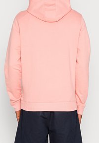 Felpa con cappuccio di un delicato rosa, con una texture accogliente. Presenta maniche lunghe e orlo a costine. La vista posteriore mostra un design semplice senza stampe.