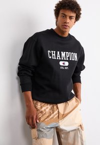 Hanorac negru cu litere și logo alb "CHAMPION"; are manșete întărite. Asortat cu pantaloni cargo bej și gri cu un model.