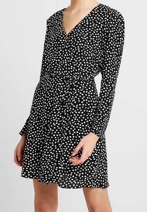 Vestido cruzado negro con lunares blancos, mangas largas, escote en V y detalle de lazo lateral, confeccionado con tela ligera.