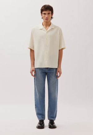 Young man wearing a cream short-sleeve button-up shirt, blue jeans, and black dress shoes standing against a plain light background.