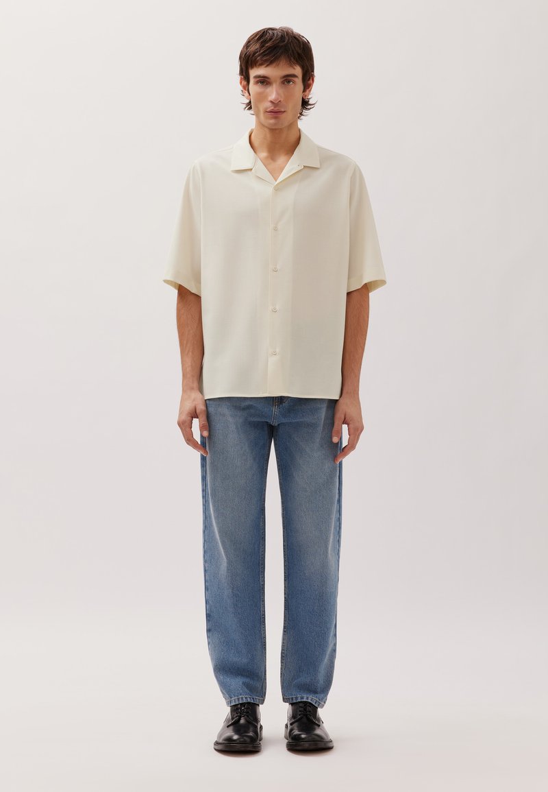 Junger Mann, der ein cremefarbenes kurzärmeliges Hemd mit Knöpfen, blaue Jeans und schwarze elegante Schuhe trägt und vor einem schlichten hellen Hintergrund steht.