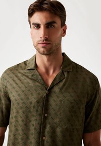 Chemise à manches courtes en tissu vert foncé, présentant un motif géométrique subtil, un col en V et une petite poche poitrine avec des boutons marron.