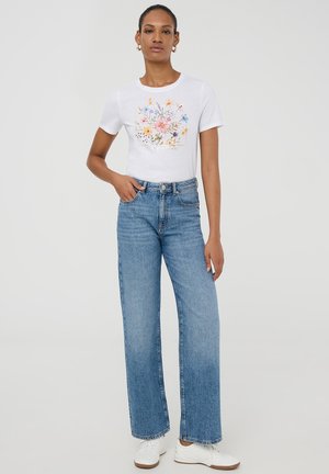 T-shirt bianca in cotone con stampa floreale e testo, abbinata a jeans in denim azzurri a vita alta e sneakers bianche.