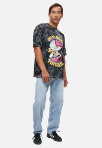 Maglietta nera con effetto tie-dye e grafica colorata; indossata con jeans azzurri chiari e sneakers nere. Vestibilità casual e rilassata.