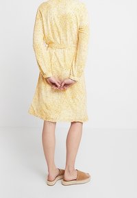 Žluté šaty po kolena s květinovým vzorem, dlouhými rukávy, zúženým pasem a detailem na zavazování na zádech. Nosit s béžovými nazouvacími sandály.