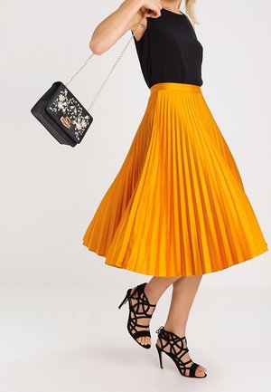 Kobieta w czarnej bezrękawniku i jasnopomarańczowej plisowanej spódnicy midi, niosąca czarną torebkę w kwiaty, ubrane w czarne sandały na wysokim obcasie z paskami.