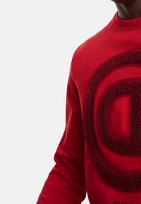 Suéter rojo con cuello redondo y mangas largas, que presenta un gran logo circular texturizado en tela de rizo en contraste.