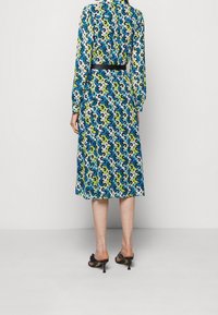Robe à motifs floraux en bleu, vert, jaune et blanc ; manches longues, longueur midi, avec une ceinture noire à la taille. Portée avec des sandales à talons noires.
