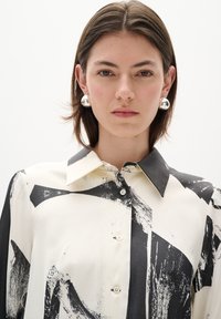 Black and white patterned shirt with a pointed collar, button front, and long sleeves made of smooth fabric. Silver circular earrings featured.