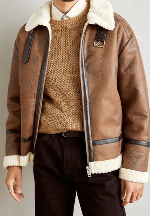 Uomo che indossa una giacca marrone in montone con colletto e polsini in pile bianco, maglione beige lavorato a maglia, camicia bianca con colletto e pantaloni scuri.