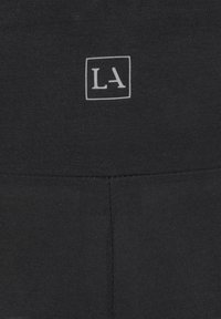 Crne tajice s glatkom teksturom, s bijelim pravokutnim logotipom "LA" na pojasu i ušivenim detaljima duž šavova.