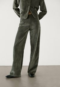 Grünes Samtanzug-Set mit einer taillierten Jacke, weit geschnittenen Hosen und subtiler Satinierung. Schwarze Schuhe ergänzen die Textur und Farbe des Outfits.