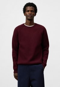 Bordeauxfarbener, gerippter Strickpullover mit Rundhalsausschnitt und Raglanärmeln, kombiniert mit navyfarbener Hose. Weiche Textur und lockere Passform.