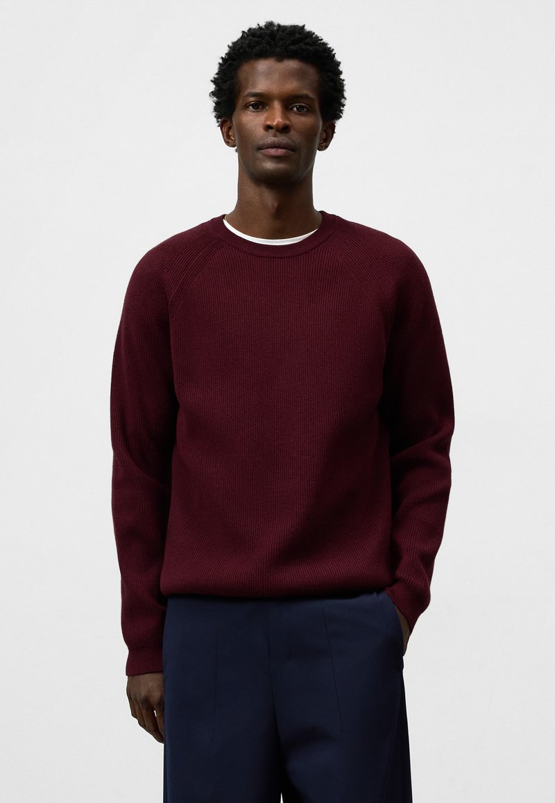 Bordeauxfarbener, gerippter Strickpullover mit Rundhalsausschnitt und Raglanärmeln, kombiniert mit navyfarbener Hose. Weiche Textur und lockere Passform.