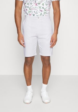 Mann trägt weiße kniehohe Shorts, weiße Socken, weiße Turnschuhe und ein Hemd mit floralem Muster mit Vögeln und Blättern.