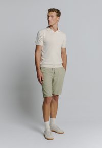 Beige polo shirt met korte mouwen, gemaakt van zachte stof; gecombineerd met lichtgroene shorts. Persoon draagt witte sokken en schoenen.