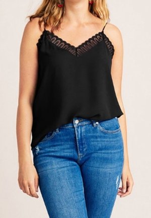 Frau trägt ein schwarzes Spitzen-Camisole-Top, in hoch taillierte blaue Jeans gesteckt, steht mit entspannten Armen an den Seiten.