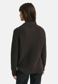 Brauner strukturierter Pullover mit einem gerippten Kragen und Bündchen, der eine lockere Passform und ein charakteristisches gewebtes Muster aufweist, von hinten betrachtet.