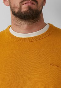 Mosterdgeel gebreide trui met een ronde hals en geribde mouwen, voorzien van een subtiele geborduurde logo op de borst. Zachte textuur.