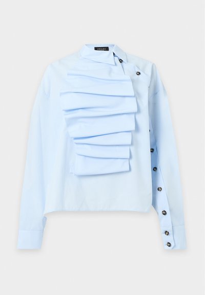 Blouse bleu clair à manches longues avec détail de volants superposés sur la poitrine et boutons noirs le long du côté droit et au niveau du poignet.