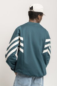 Teal sweatshirt med hvide stribe detaljer på ærmerne, med en rund halsudskæring og ribbede manchetter. Båret med lyseblå jeans.