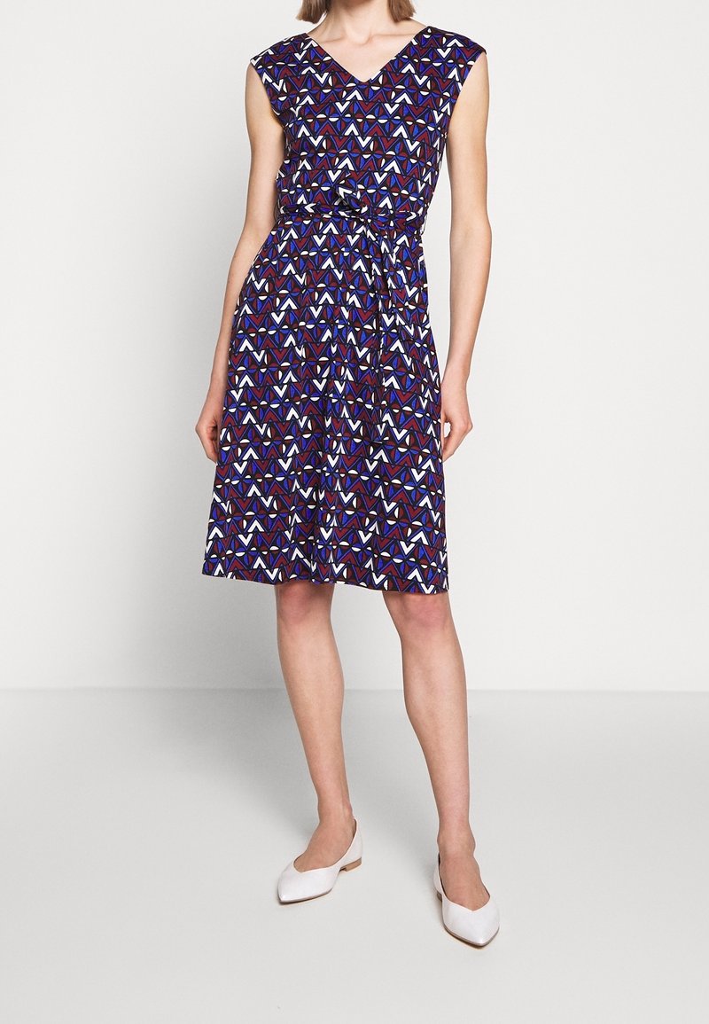 Robe sans manches, longueur genou, de couleur violet foncé, avec un motif géométrique rouge, blanc et bleu. Ceinture nouée à la taille, tissu lisse.