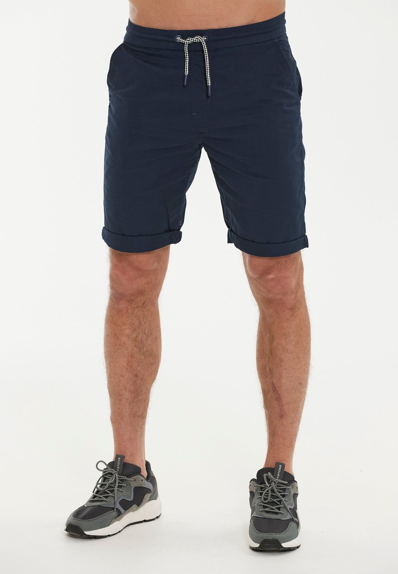 Marineblaue Shorts mit Kordelzugbund, seitlichen Taschen und umgeschlagenen Säumen. Kombiniert mit schwarz-grauen Sportschuhen. Glatte Textur.