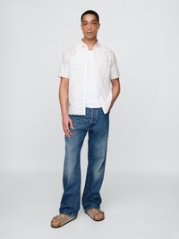 Hvit kortermet skjorte med åpenmønster, kombinert med blå denimjeans og grå sandaler. Modellen står med hendene i lommene.