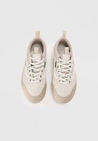 Un par de zapatillas de lona beige con cordones blancos, punteras de goma y tiradores, vistas desde arriba sobre un fondo blanco.