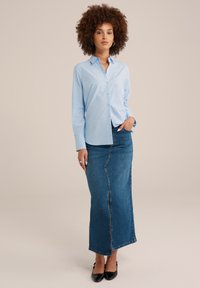 Donna con capelli ricci che indossa una camicia azzurro chiaro con bottoni, una lunga gonna di jeans e tacchi neri, in piedi con una mano in tasca su uno sfondo beige.